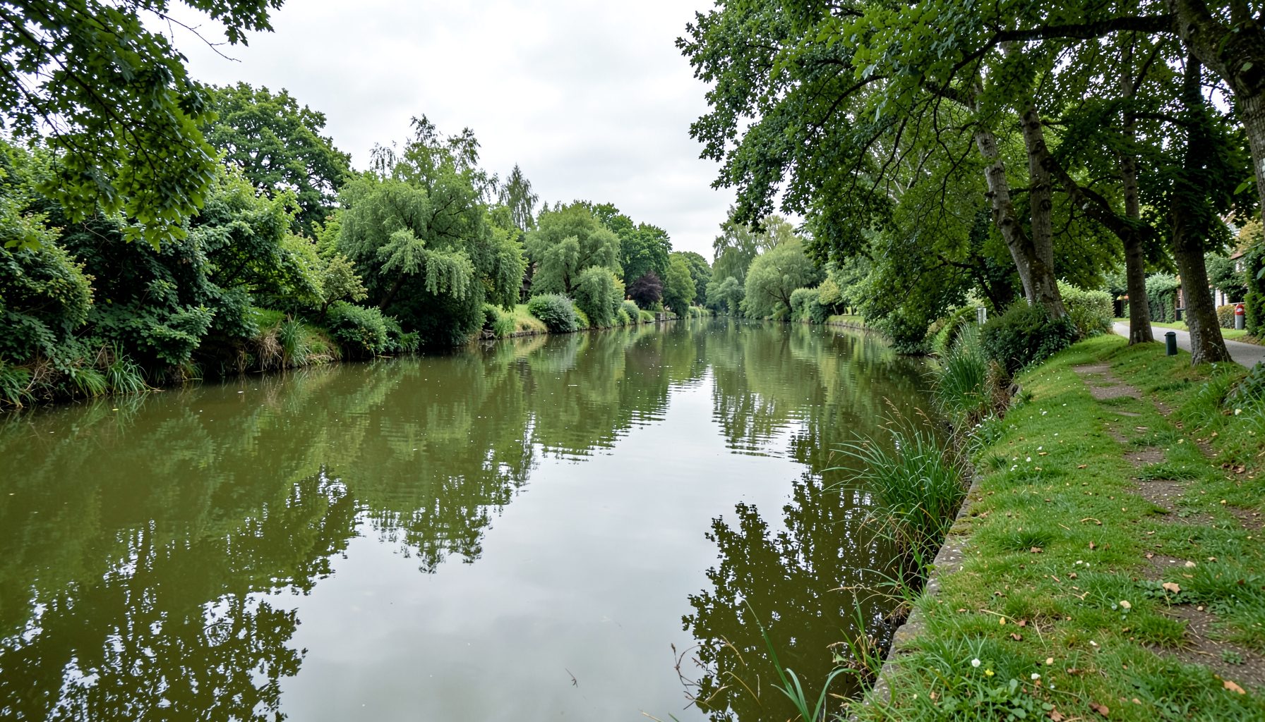 A tranquil English canal scene with still green-brown water reflecting overhanging trees and a grassy towpath stretching into the distance under soft grey skies